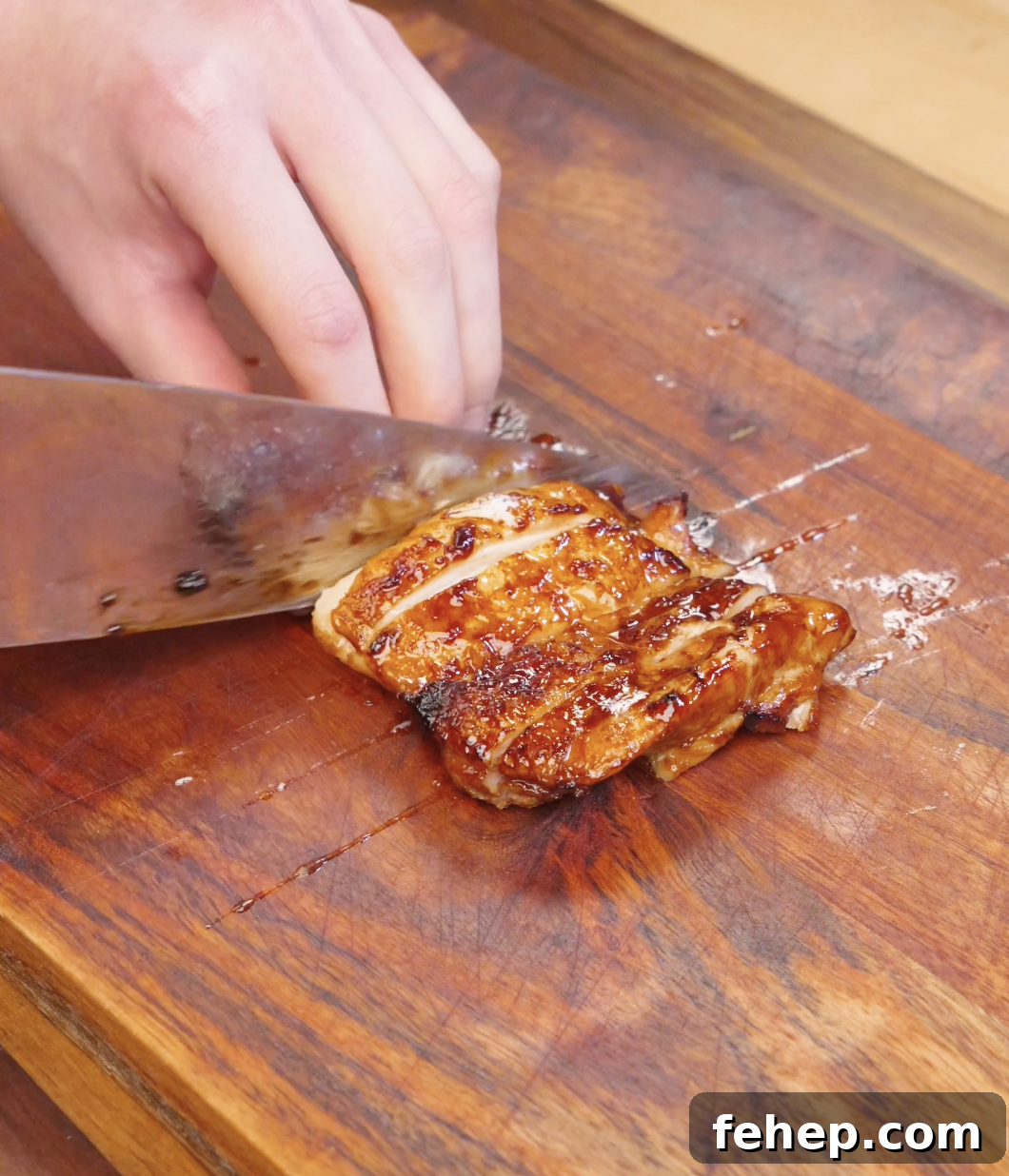 Knife slicing cooked Sweet Chili Chicken thighs into strips on a wooden cutting board.