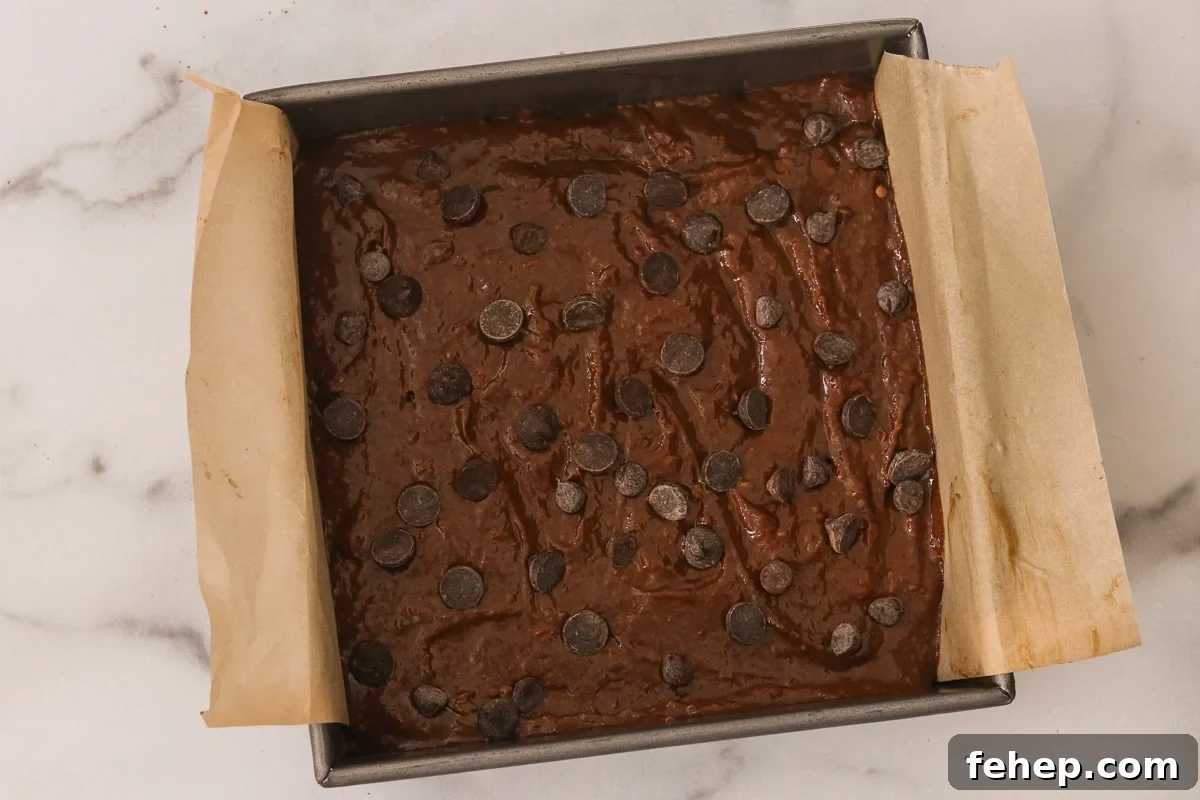 Sweet potato brownie batter in a parchment-lined pan spread out evenly. Remaining chocolate chips are sprinkled over the top.