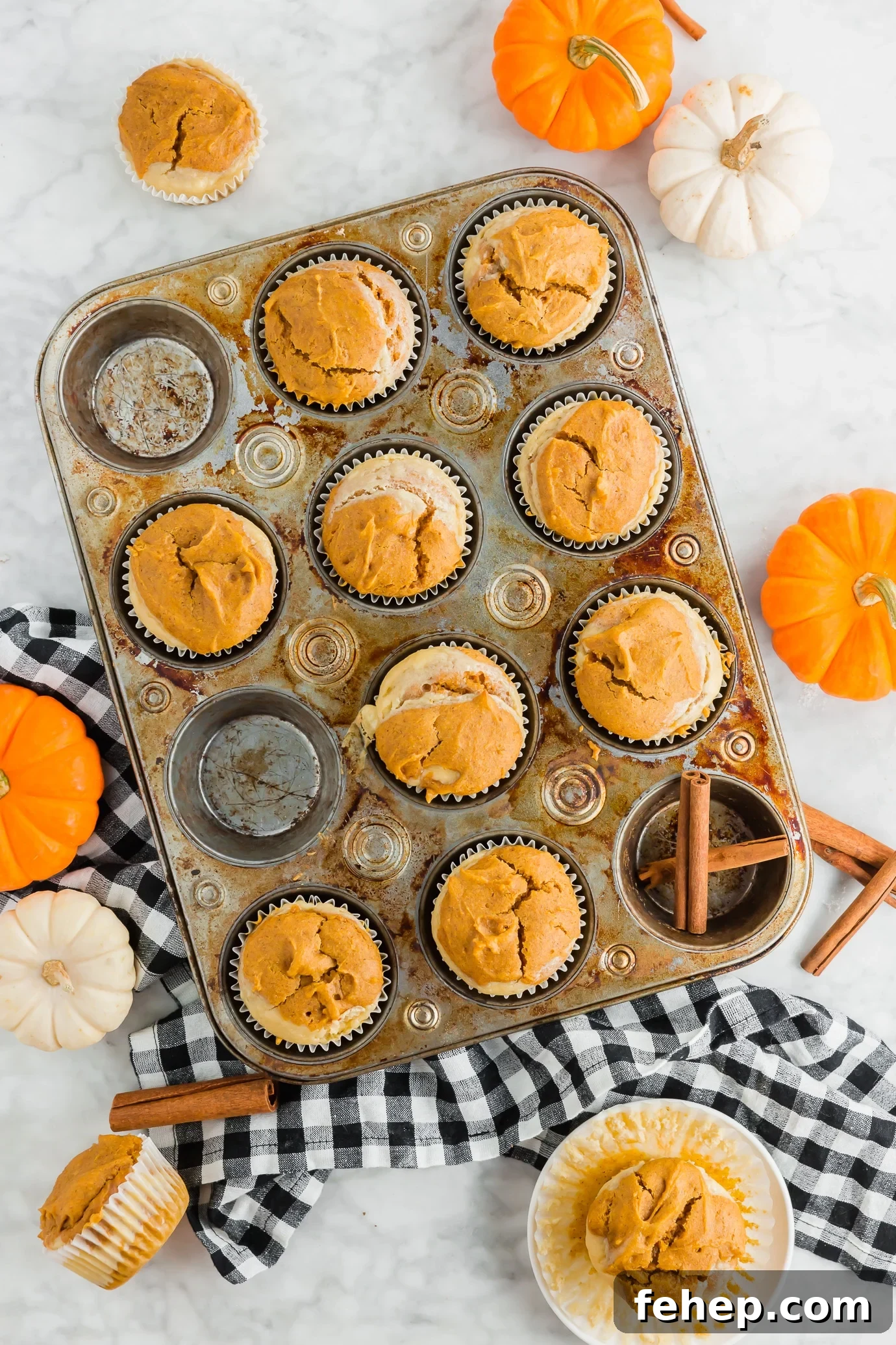 Gluten-Free Pumpkin Cream Cheese Muffins A photo of a muffin tin filled with gluten-free pumpkin cream cheese muffins and cinnamon sticks and mini pumpkins.