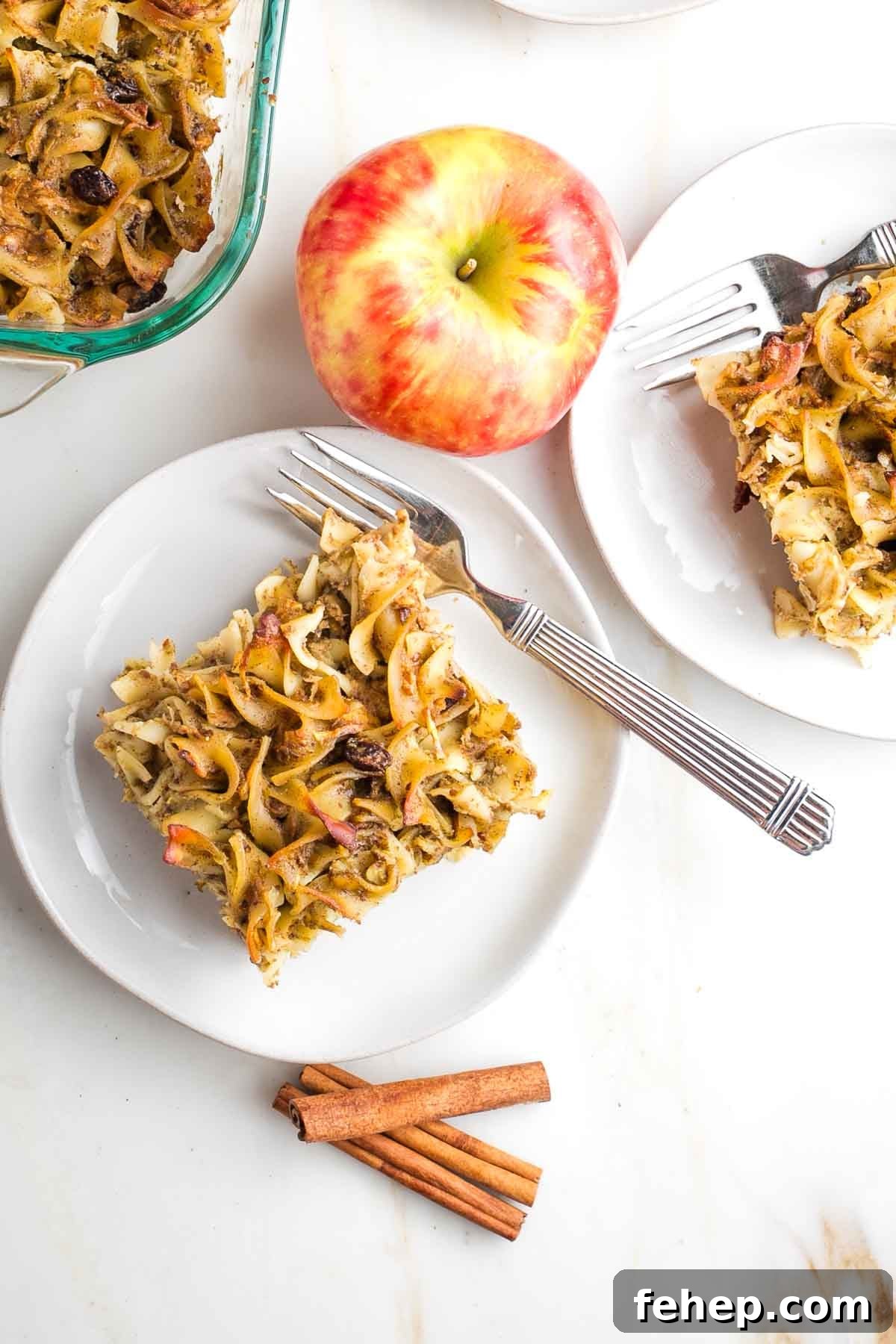 Piece of noodle kugel with raisins on a plate with cinnamon sticks and an apple in the background.