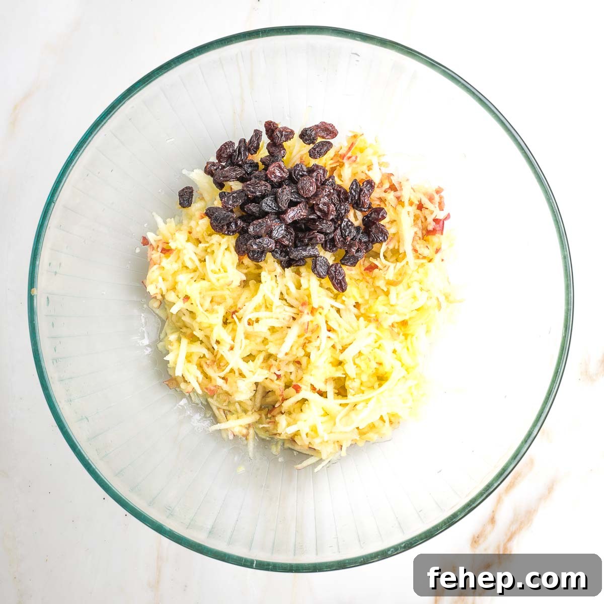 Shredded apples and dark raisins in a large glass mixing bowl. 