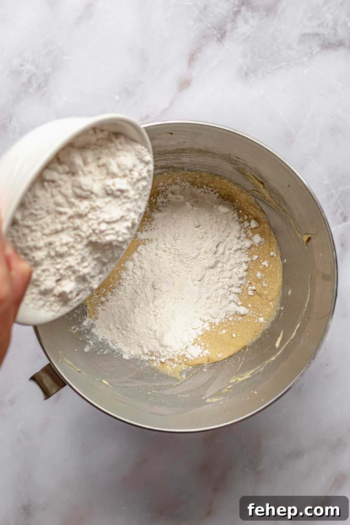 A hand pours a bowl of dry ingredients into wet ingredients.
