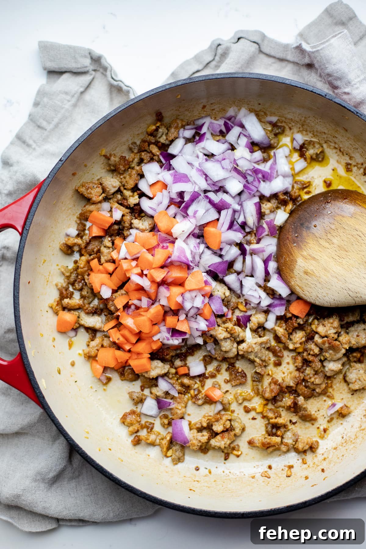 Veggies and sausage being cooked in a red pan with a wooden spoon.