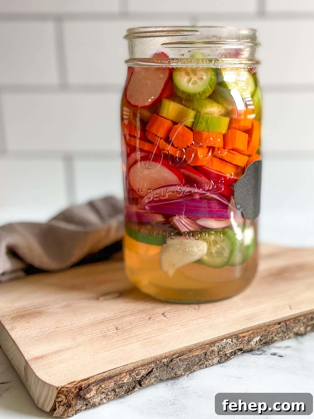 A glass jar of mixed pickles sits on a wooden cutting board with a gray linen.