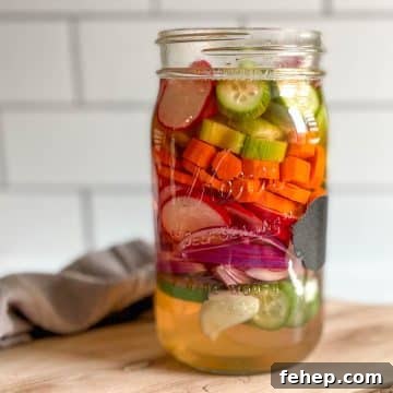 A glass jar of mixed pickles sits on a wooden cutting board with a gray linen.