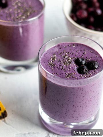 Two small glasses with a purple smoothie topped with blueberries with a bowl of blueberry in the back and a banana.