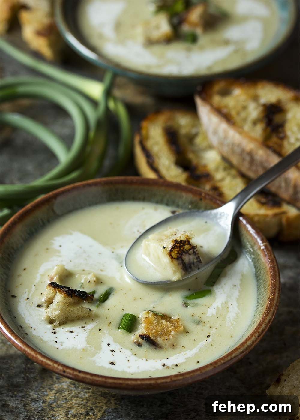 A spoonful of garlic scape soup with a crouton. Another bowl of soup, grilled bread, and garlic scapes in the background.
