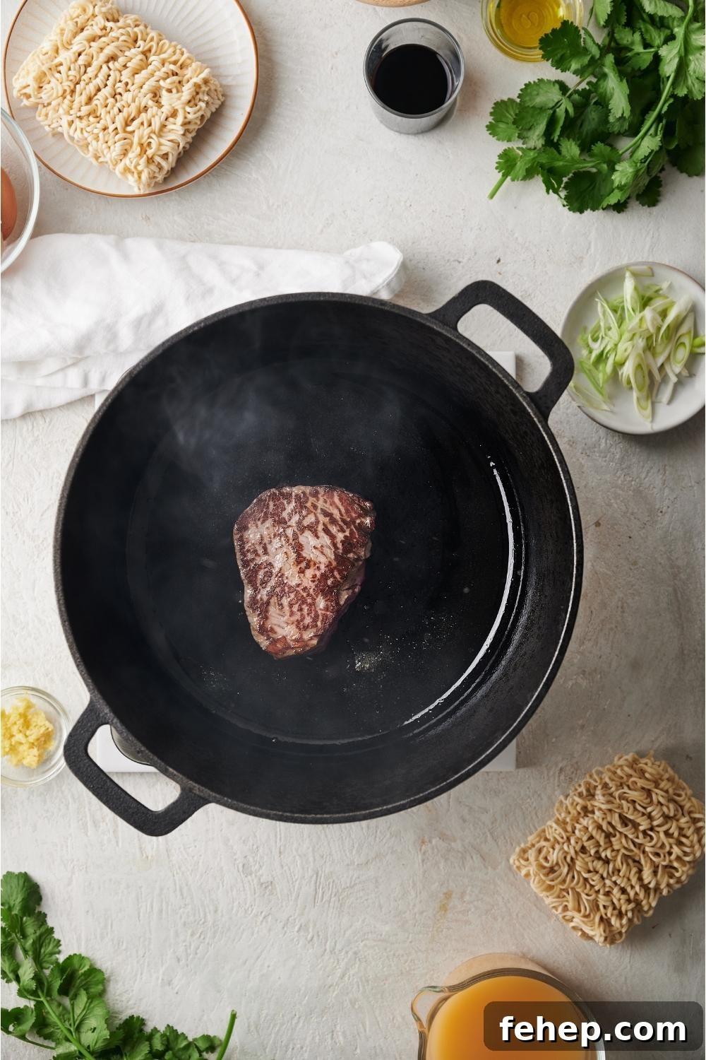 Black dutch oven with sirloin searing in the pan, surrounded by ingredients to make beef ramen